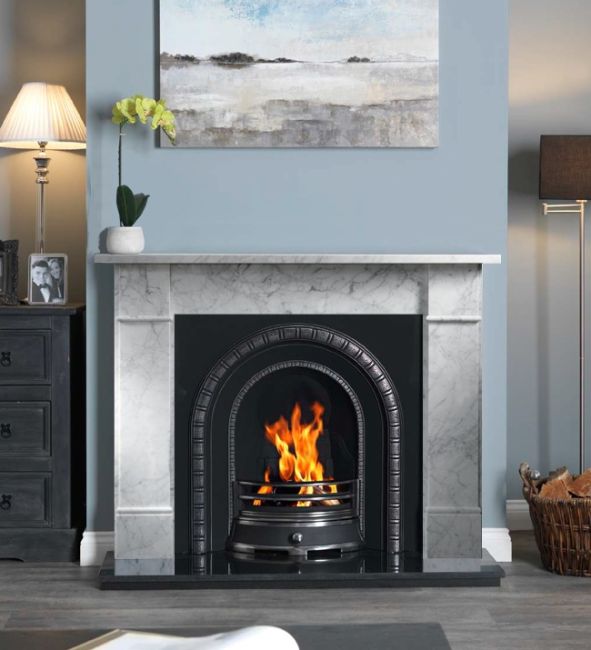 Brompton carrara marble foireplace surround with a cast iron insert and a roaring fire. On a light blue wall with a seascape canvas above the fireplace. There is grey carpet on the floor, a basket filled with logs to one side. In one alcove there is a floor lamp, in the other, a sideboard with a table lamp switched on. 