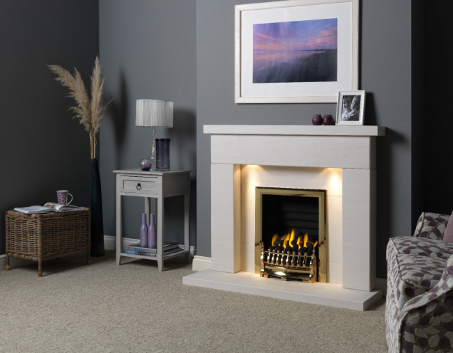 The Gallery Collection Durrington Limestone Fireplace surround in white featuring the Blenheim gas fire with a gold trim. The fire surround has warm dwonlights and a white back panel and hearth. In the room, there are grey walls and a beige carpet. There is a framed image of a nightscape with a purple sky above the fire and a framed photograph next to two candle holders on the mantel shelf. In the alcove, there is a small grey side table with a white lamp, and decorative pampas grass in a vase next to a wicker basket with a lid that has a mug and an open book on top. 