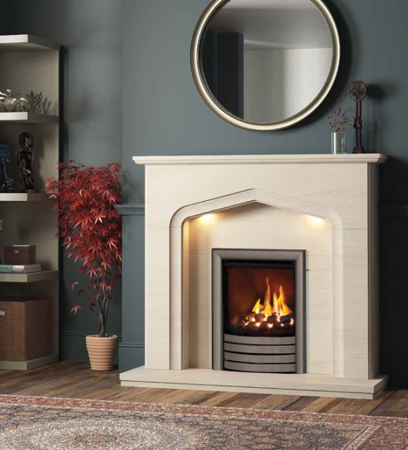 The Elign & Hall Aurelia limestone fireplace surround in cream. The surround has a tudor arch and warm downlights. The back panel and hearth matches the surround and it has an inset fire with a silver trim. In the room, the walls are a blue-grey and there is a round mirror hanging above the fireplace. On the mantel, there is a small vase and candle holder. The floor is wooden and there is a floral rug. A pot with a red leafy plant is sitting next to the fireplace. In the alcove, there are modern, slim shelves filled with books and decorative items. 