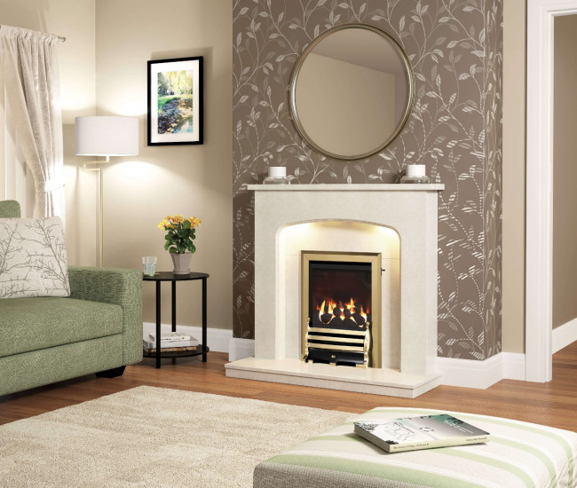 The Flare Tasmin Micro Marble fireplace surround in cream with a brass trim gas fire and downlights. The wall has a floral brown wallpaper and there is a round mirror hanging above the fireplace. There is a green sofa, a white floor lamp, and a small painting of nature hanging on the wall in the alcove. There are two candles on each side of the mantel. The floor is wooden with a cream rug. There is a small table with a vase of yellow flowers to the side of the fireplace. 