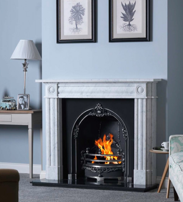 Gallery collection chiswick carrara marble fireplace surround in white with column detailing and corbels. On a light blue wall with two drawings of frees framed above the fireplace. The fire surround has a cast iron victorian style insert and a roaring fire. In the alcove, there is a console table with a framed photograph and a lamp with a white shade. To the side there is a floral armchair with a side table that has a mug on it. The carpet is grey. 