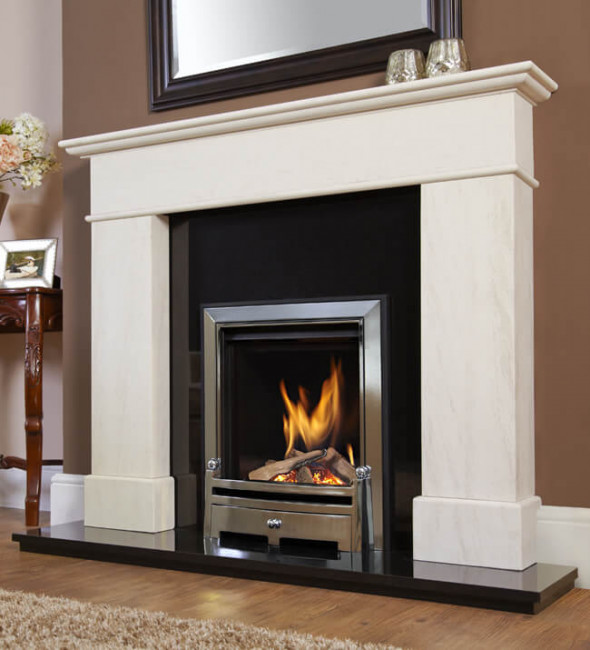 The Axon Verine Renaissance Limestone Fire surround with a black back panel and hearth, and the Verine Passion inset gas fire with a silver trim. There are brown walls, a brown wooden floor and a framed mirror hanging above the fireplace. To the side there is a sideboard with a framed photo and a vase of flowers. Two silver candle holders are sitting together on the mantel. 