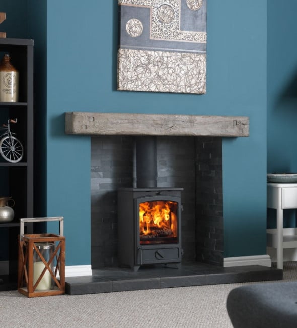 Go Eco Plus stove in a fireplace opening with a wooden beam against a blue wall. Featuring a decorative lantern, black hearth, grey carpet, and an abstract canvas painting in blue and grey hanging on the chimney breast. Black shelves with decorative items including a vase and bicycle ornament in the alcove. .